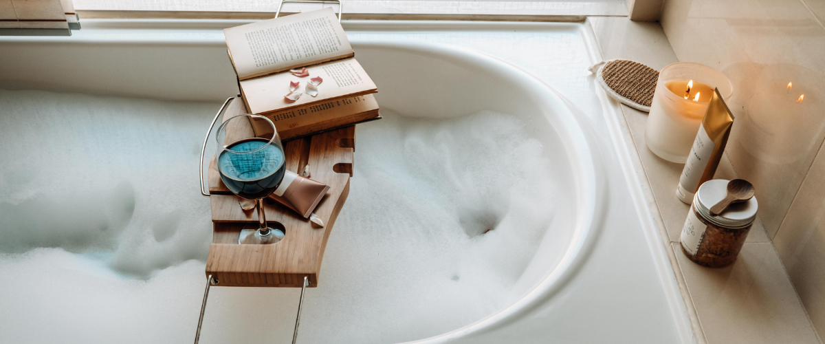 A bubble bath surrounded by candles and a bath tray with a glass of red wine and a stack of books