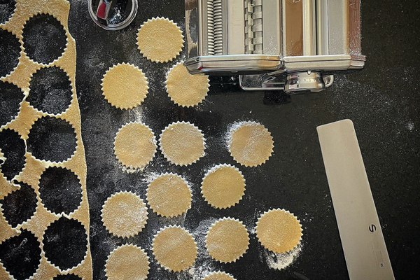 Overhead view of raw pasta in various shapes, four sprinkled across a black counter, a stainless steel pasta machine on the side