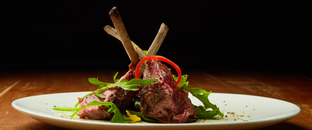 side view of a white plate on a dark background; lamb chops standing upright on a bed of lettuce leaves