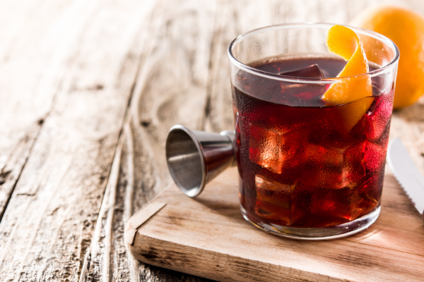 side view of a red-brown boulevardier cocktail in a rocks glass with an orange twist on a wood board