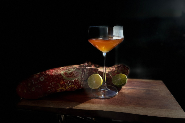an orange cocktail in a coup against a black background; a halved lime and autumn napkin beside the glass