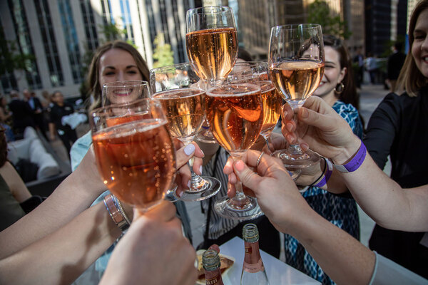 8 glasses filled with rose clinking together, with blurred smiling faces in the background
