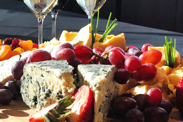 Two glasses of white wine and one of red behind an overflowing cheeseboard filled with different cheeses, grapes and strawberries