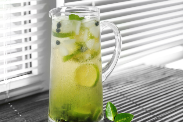 a glass pitcher filled with clear green liquid, limes and herbs