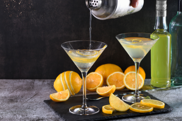 two martini glasses, one with a cocktail pouring mid stream into it. A silver shaker and pile of lemons in the background