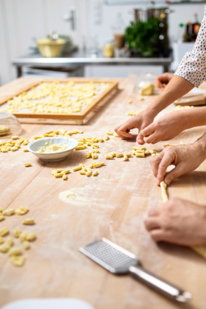 Close up on wooden table filled with pasta dough and shapes; a pair of male and female hands actively rolling dough